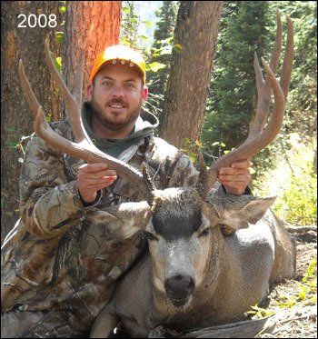 Man in camouflage and orange hat, holding large deer antlers, next to the deer in a forest, 2008.