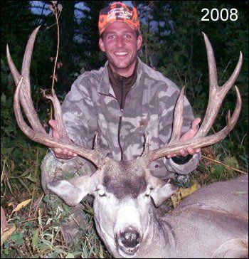 Man in camouflage holds large deer antlers, smiling in a forest. Marked as 2008.