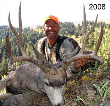 Man in camouflage holds large buck with antlers in mountain setting.