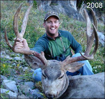Man kneels with a large buck, holding its antlers. He smiles. Outdoor setting, mountains in background. 