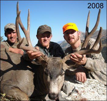 Three men hold a large deer with antlers. One wears an orange hat. Blue sky, mountains in the background. 