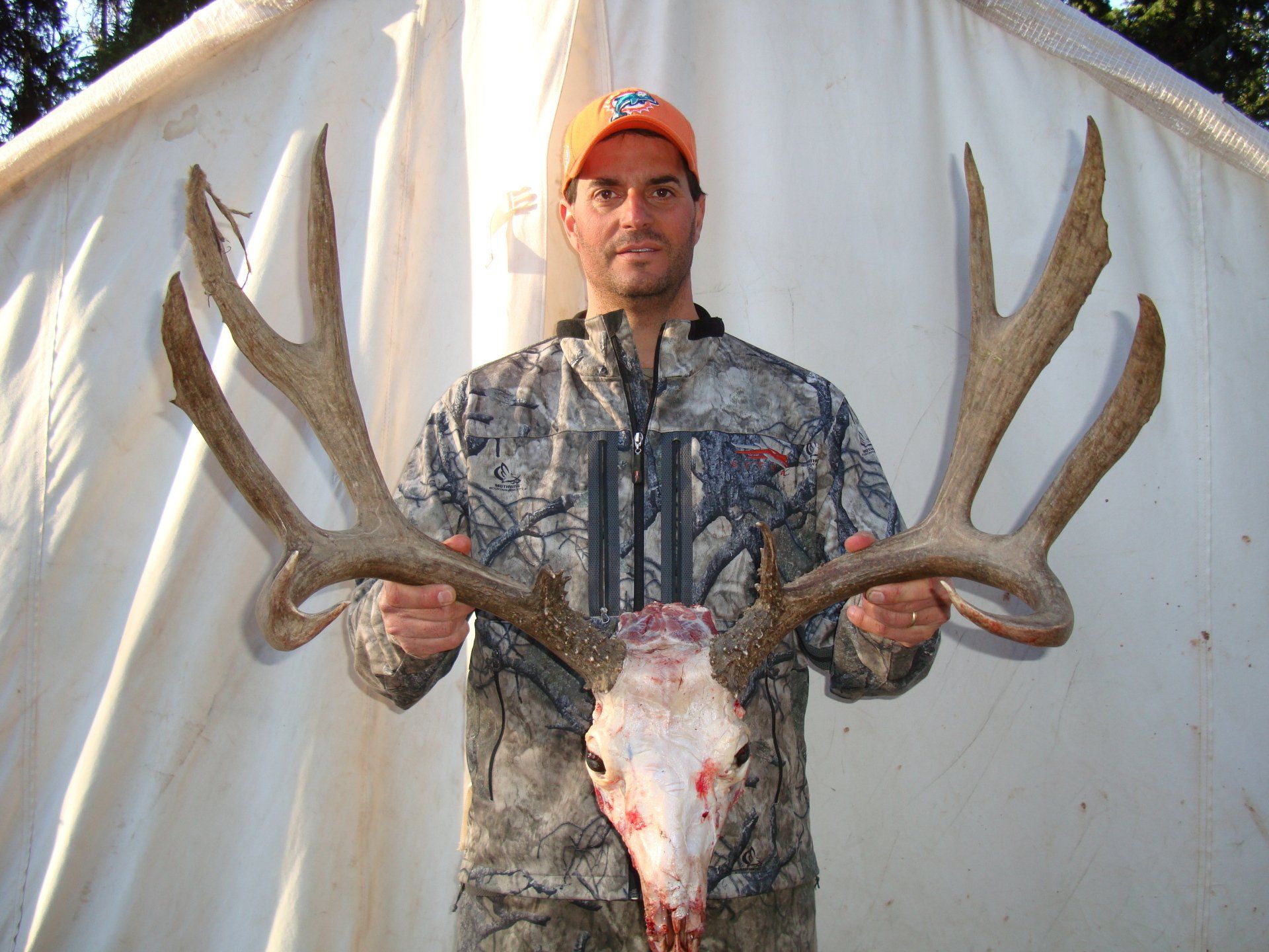 Man in camo holds deer skull with large antlers in front of a white tent.
