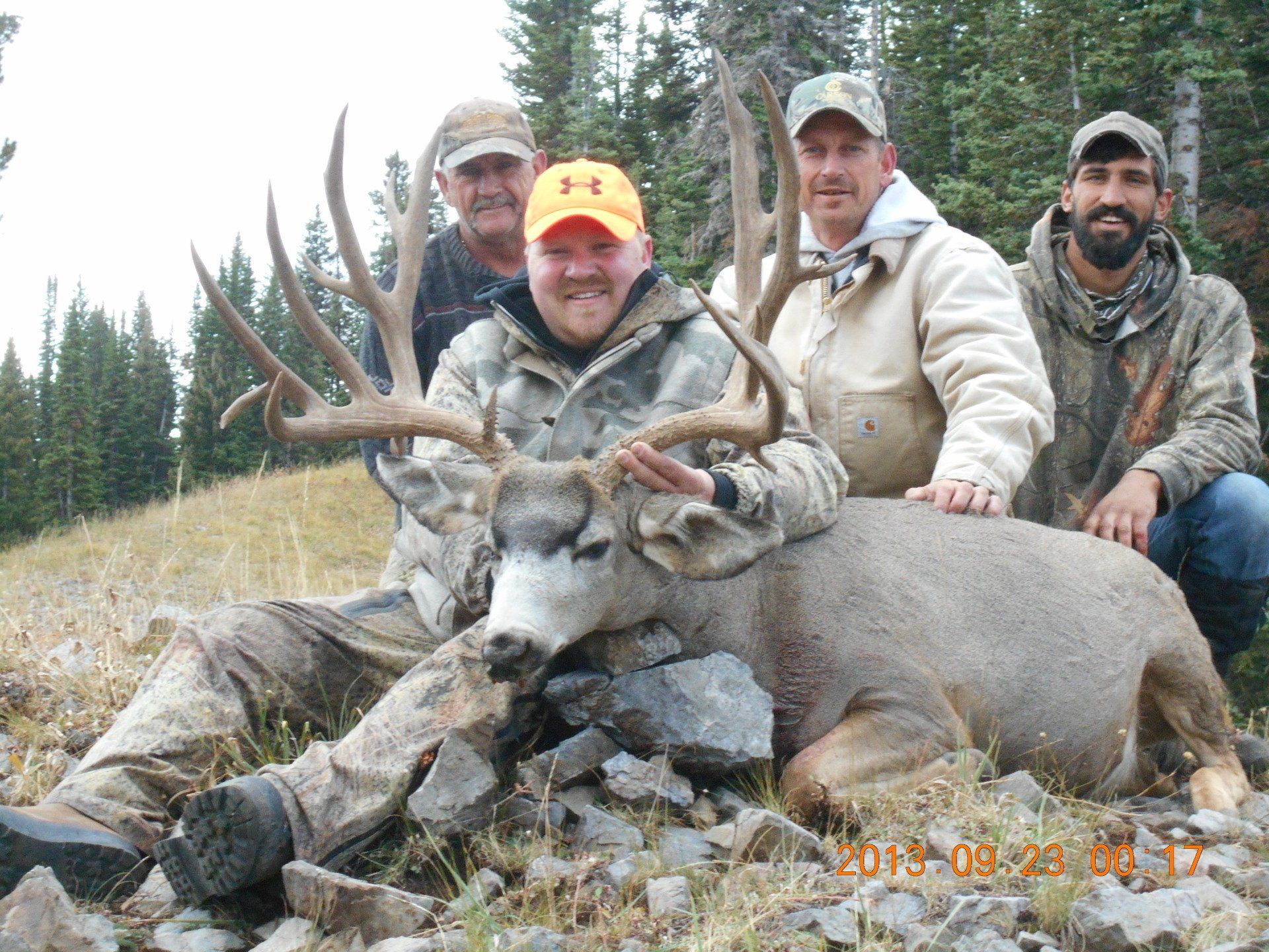 Four men pose with a large buck deer on a hillside. The buck lies on the ground with antlers. All are wearing hunting attire.