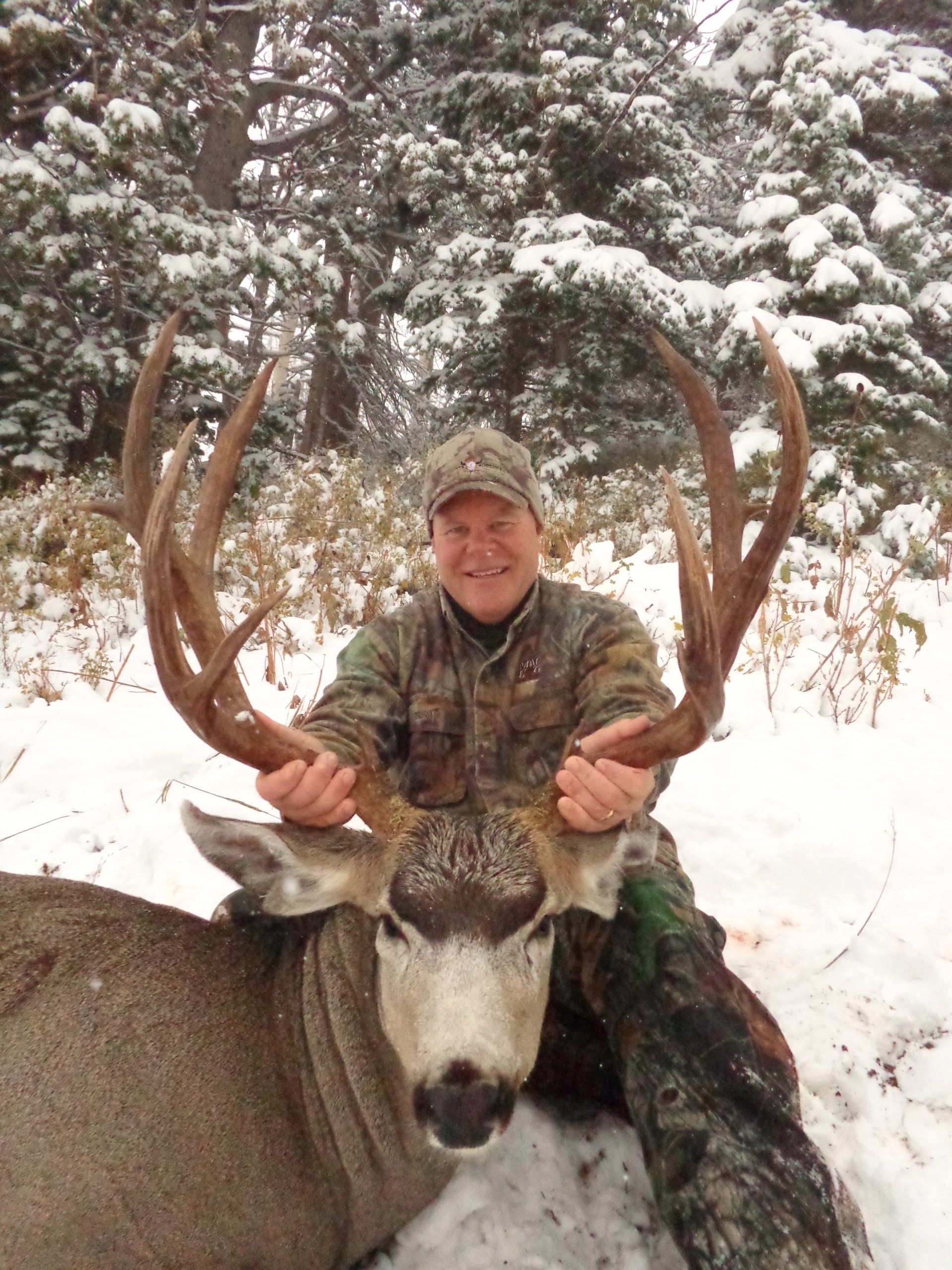Hunter in camouflage holds a large buck with impressive antlers in a snowy forest.