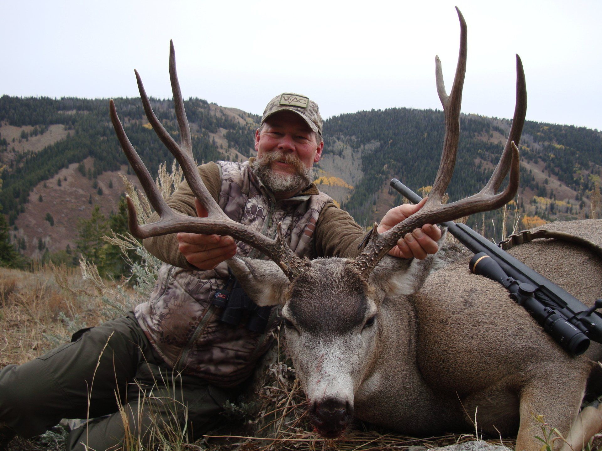 Hunter in camo holds large buck with impressive antlers outdoors; rifle visible.