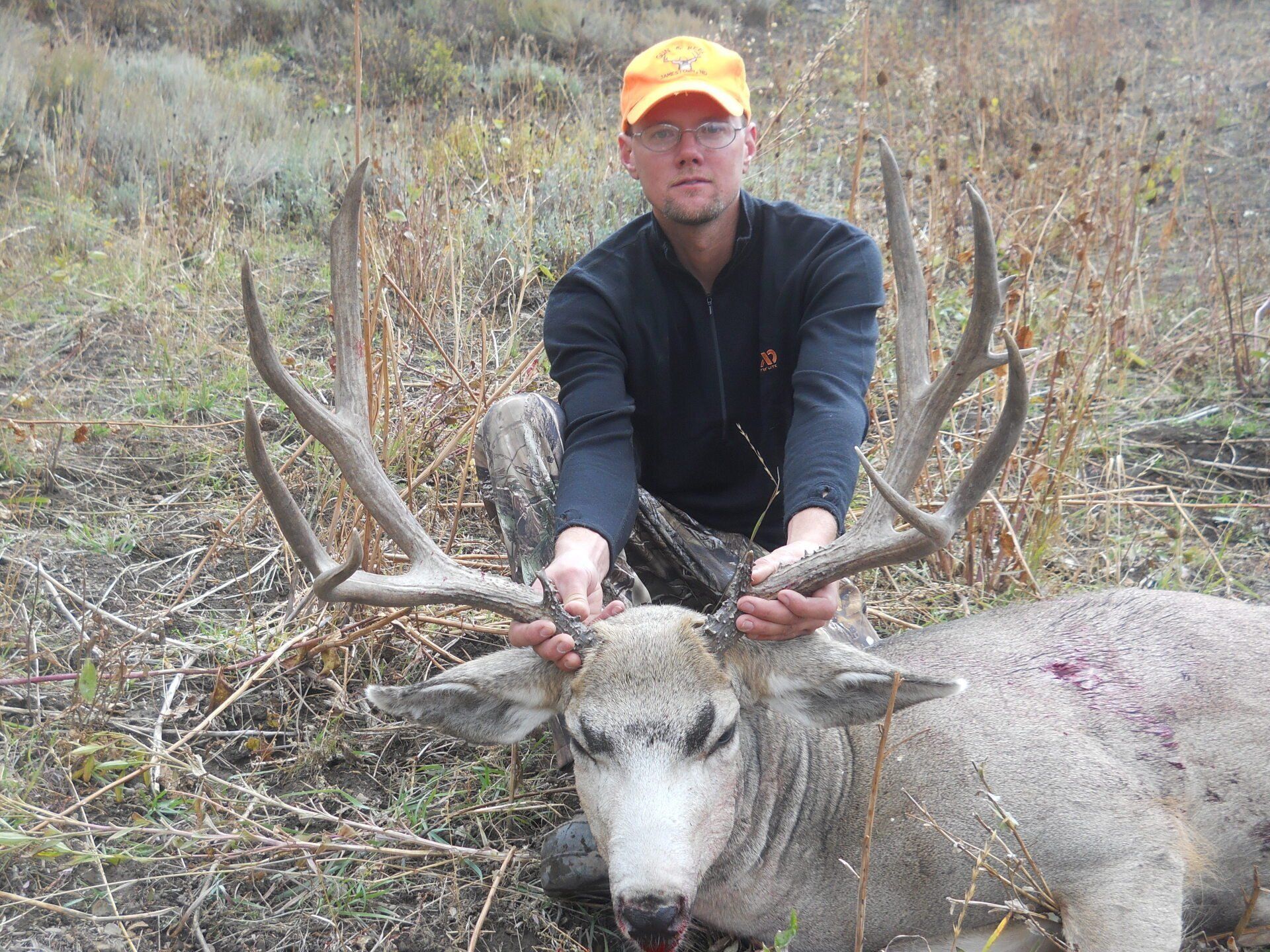 Man kneeling with a large buck. Hunter holds antlers, wearing orange cap, on grassy hillside.