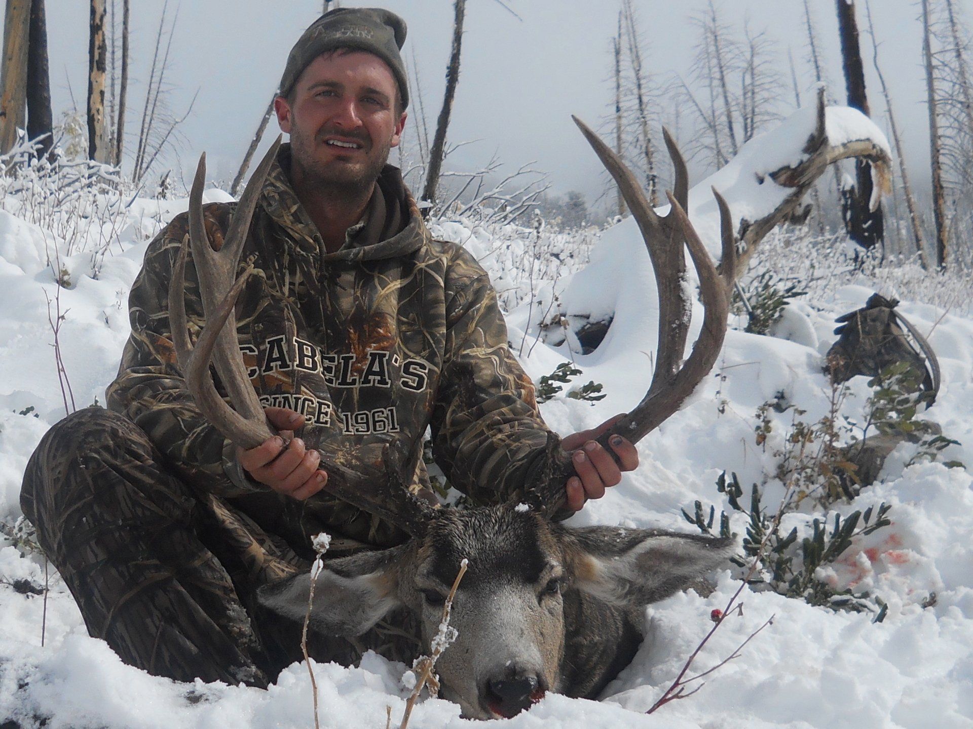 Man in camo smiles, kneeling in snow next to a large-antlered deer.