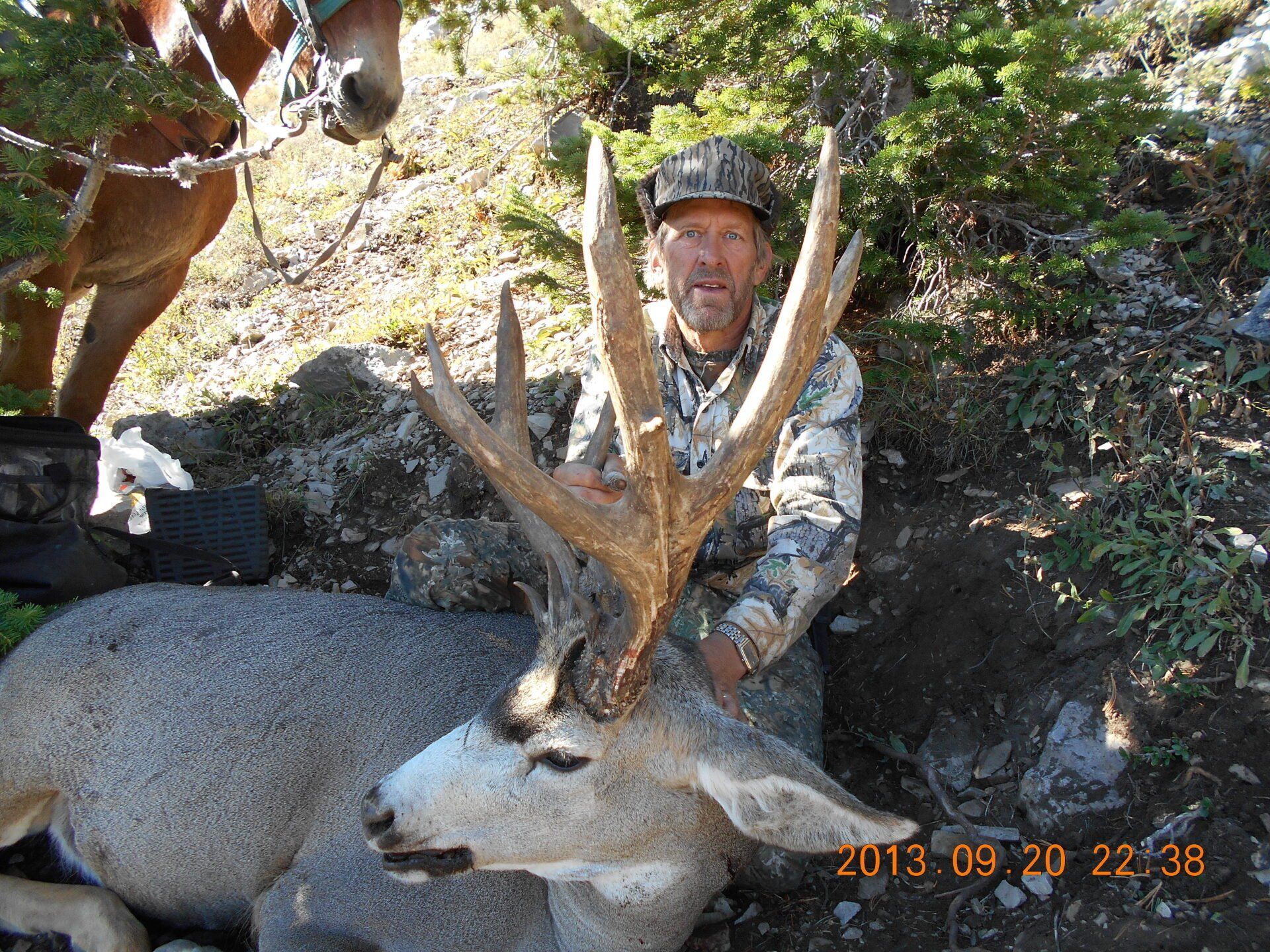 Man in camo with a large-antlered deer he has killed. The setting appears to be outdoors.