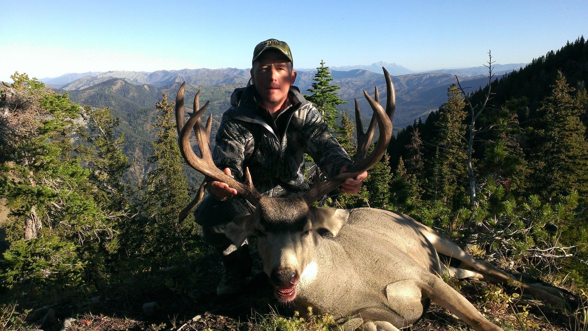 Hunter kneels beside a large buck with impressive antlers, set against a mountainous, wooded landscape.