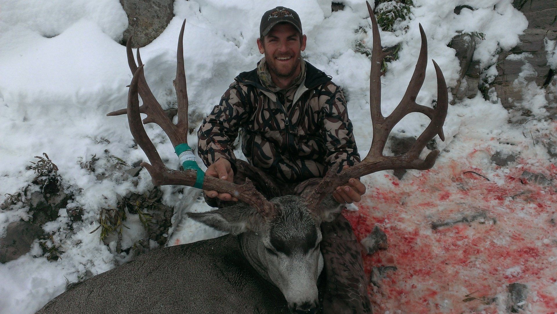 Man kneels, holding large deer antlers, with deer carcass; snow and rocks in background.
