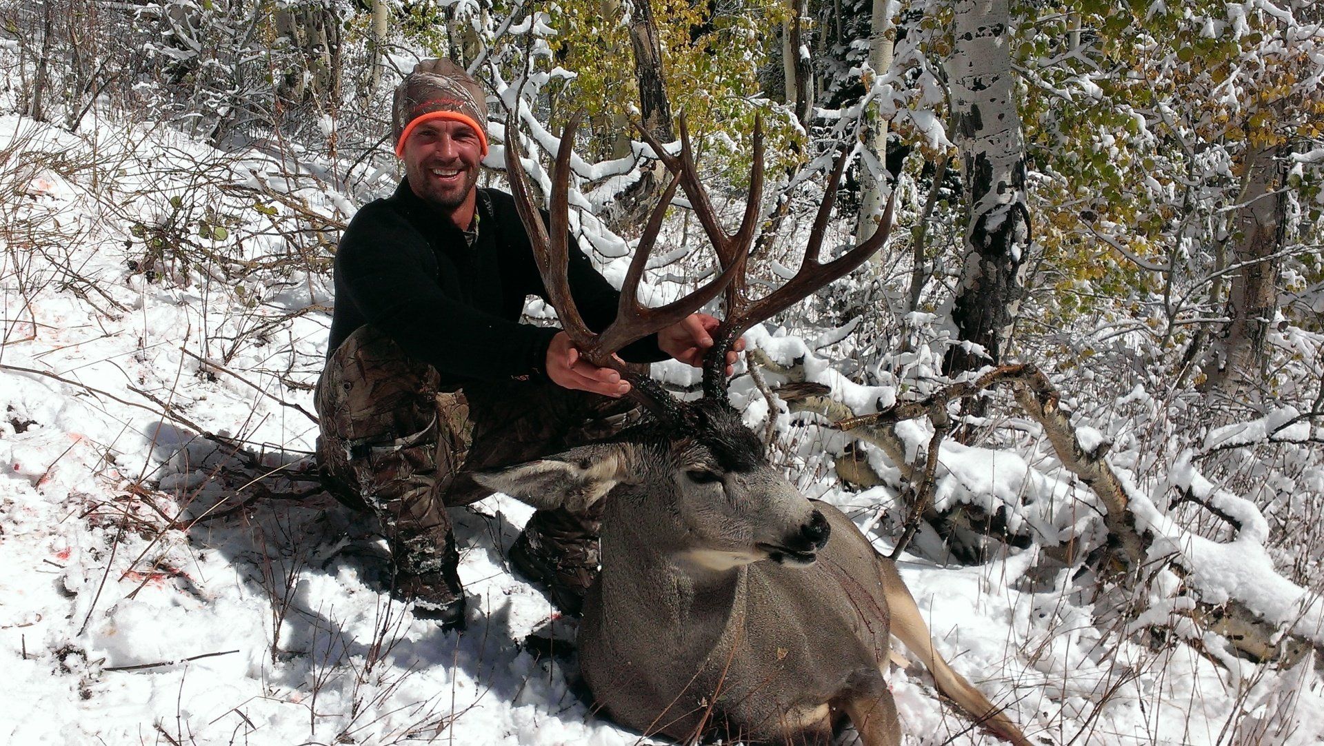 Hunter kneels with a large buck in the snow, smiling. He wears camo, orange glasses; snowy forest.