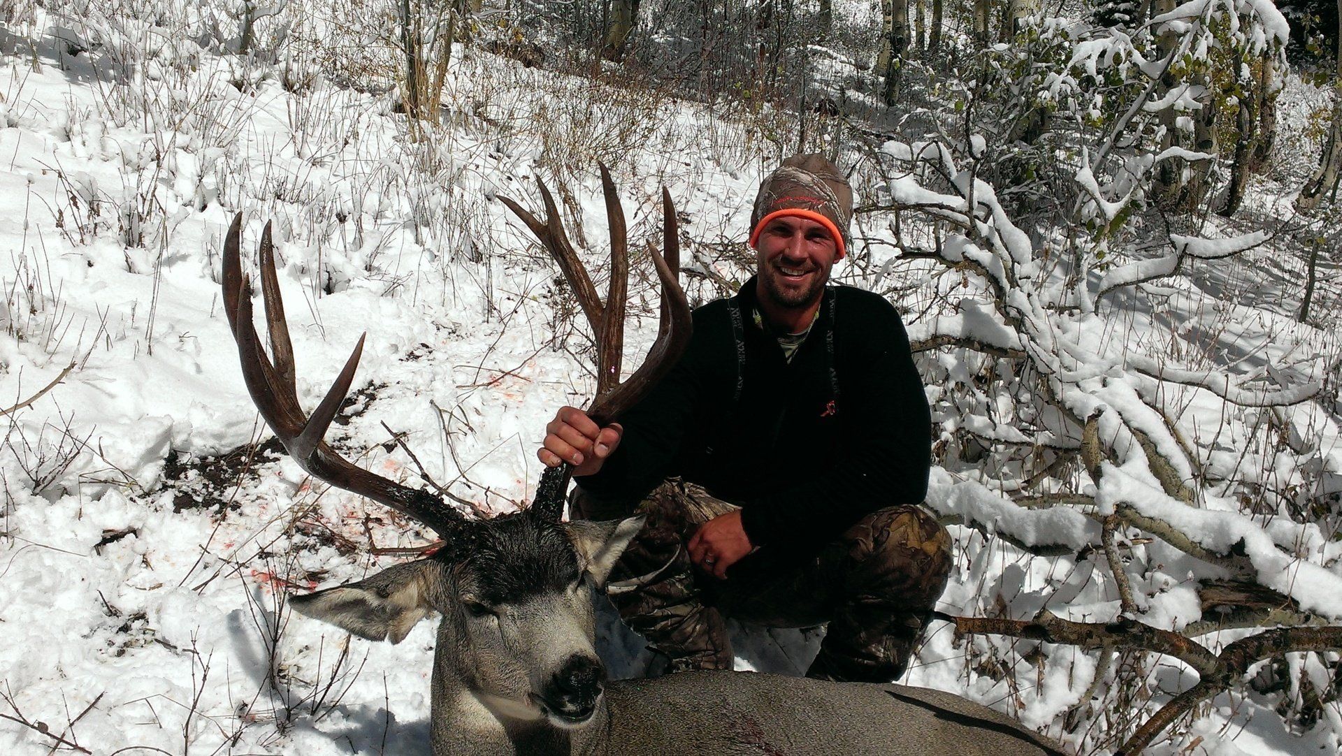 Man kneeling beside a harvested deer with large antlers in a snowy outdoor setting.