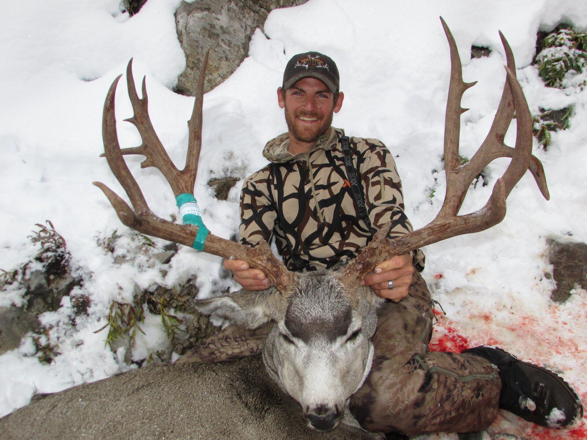 A man in a camo shirt is holding a large deer in the snow