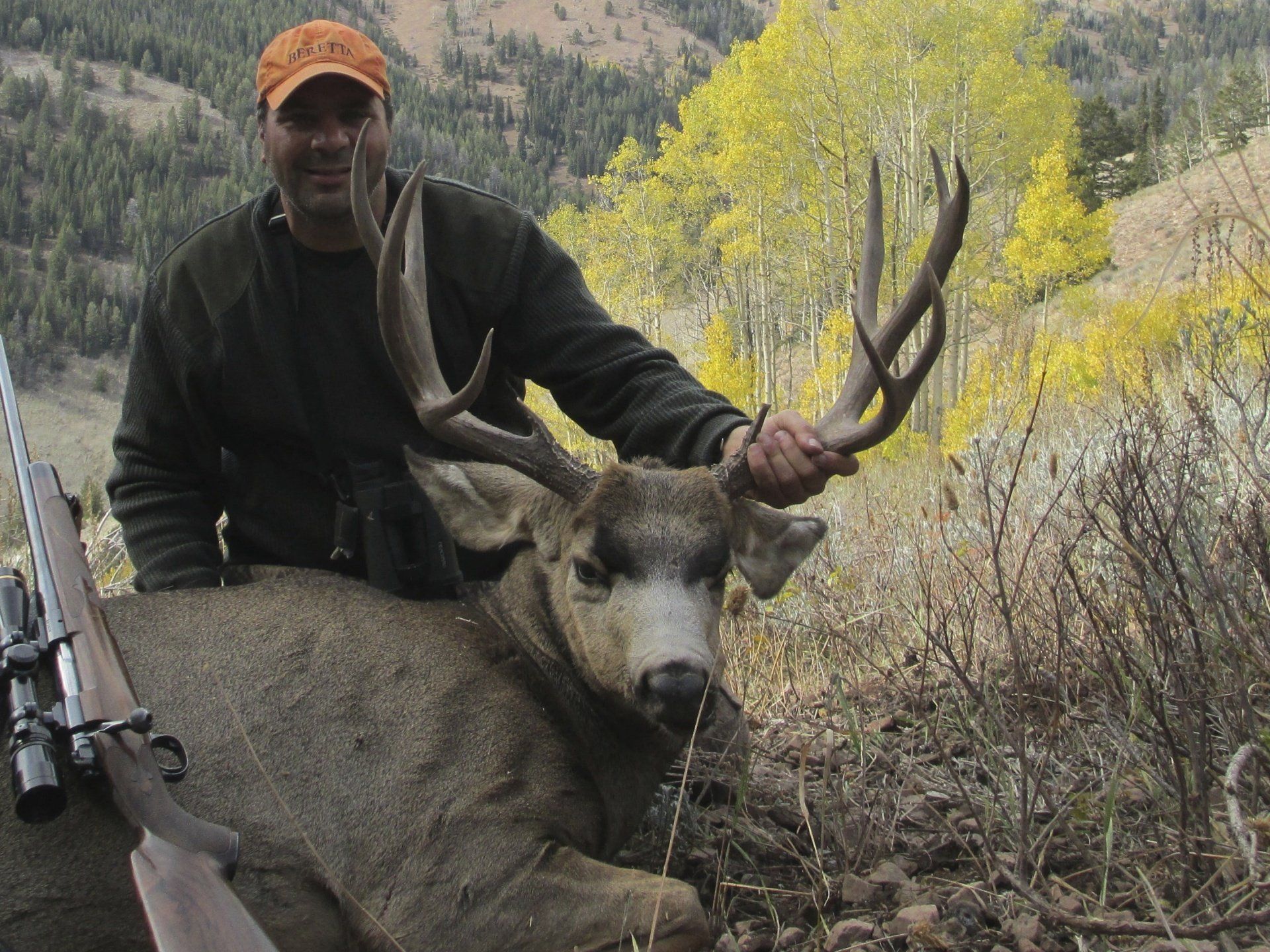 Hunter with a large buck, rifle nearby, in autumn mountain setting.