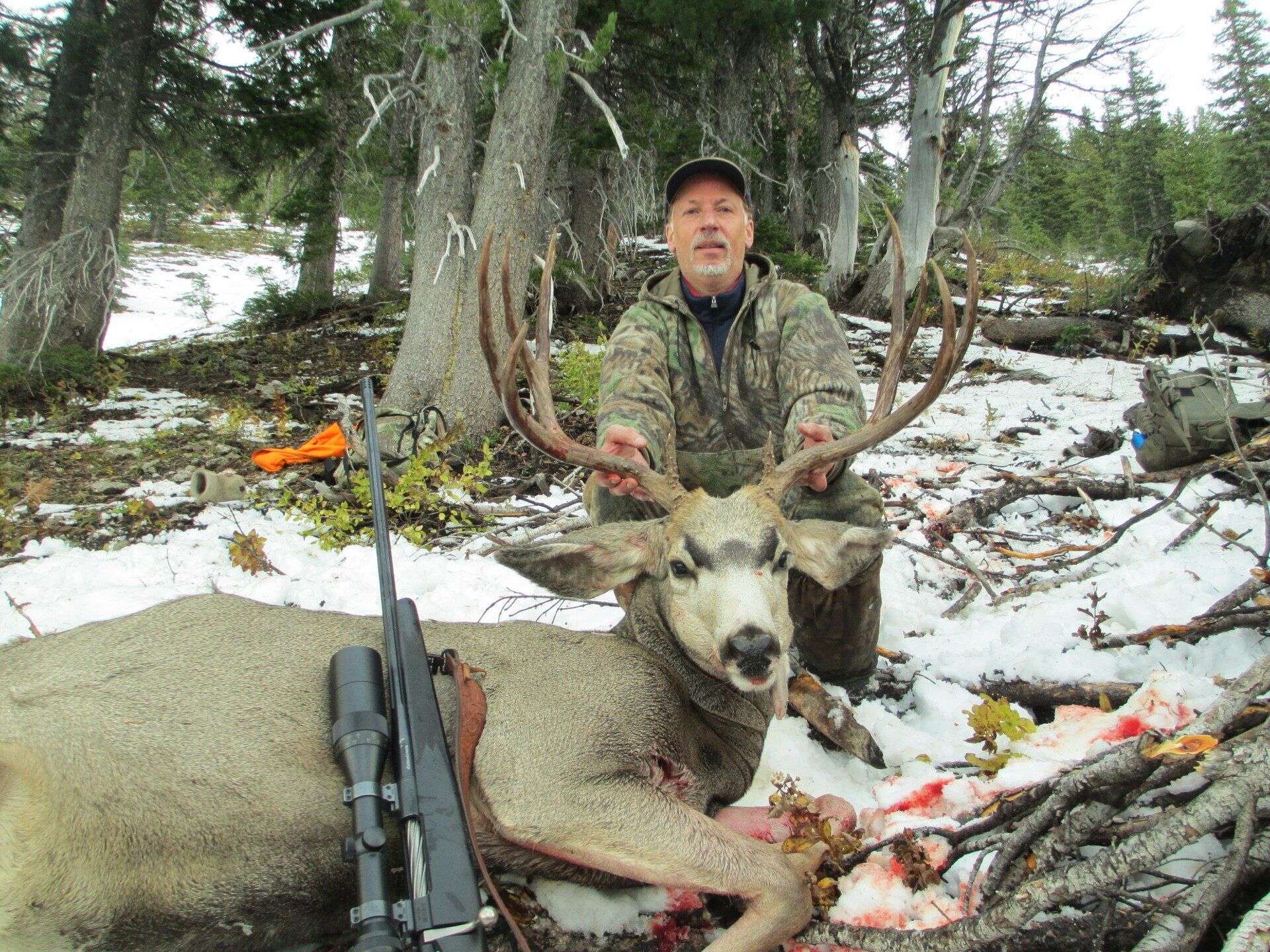 Hunter in camouflage poses with a large buck deer in a snowy forest. A rifle lies nearby.