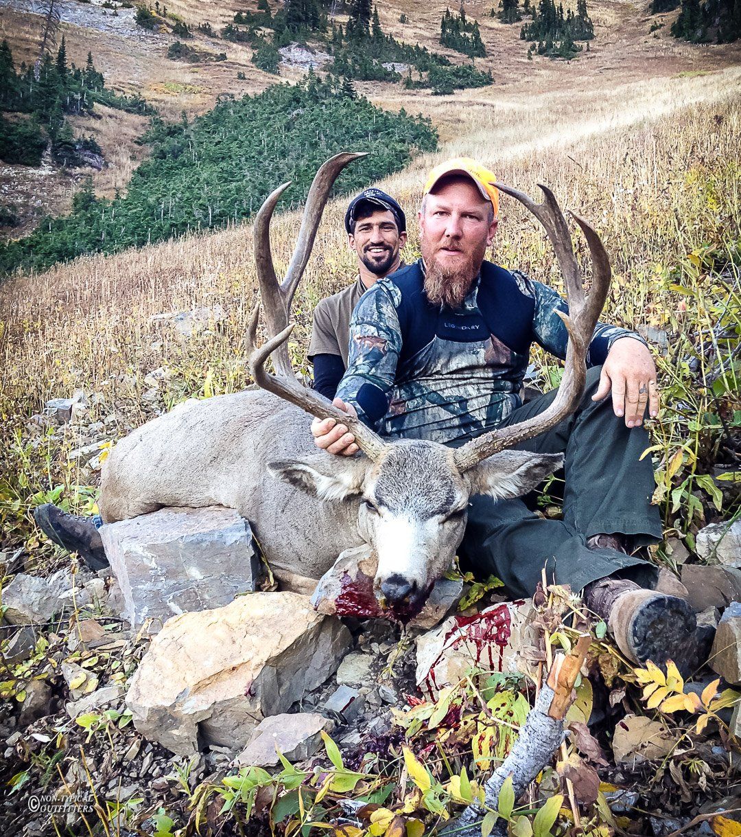 Two men pose with a dead mule deer buck in a mountainous area. One man holds the large antlers.