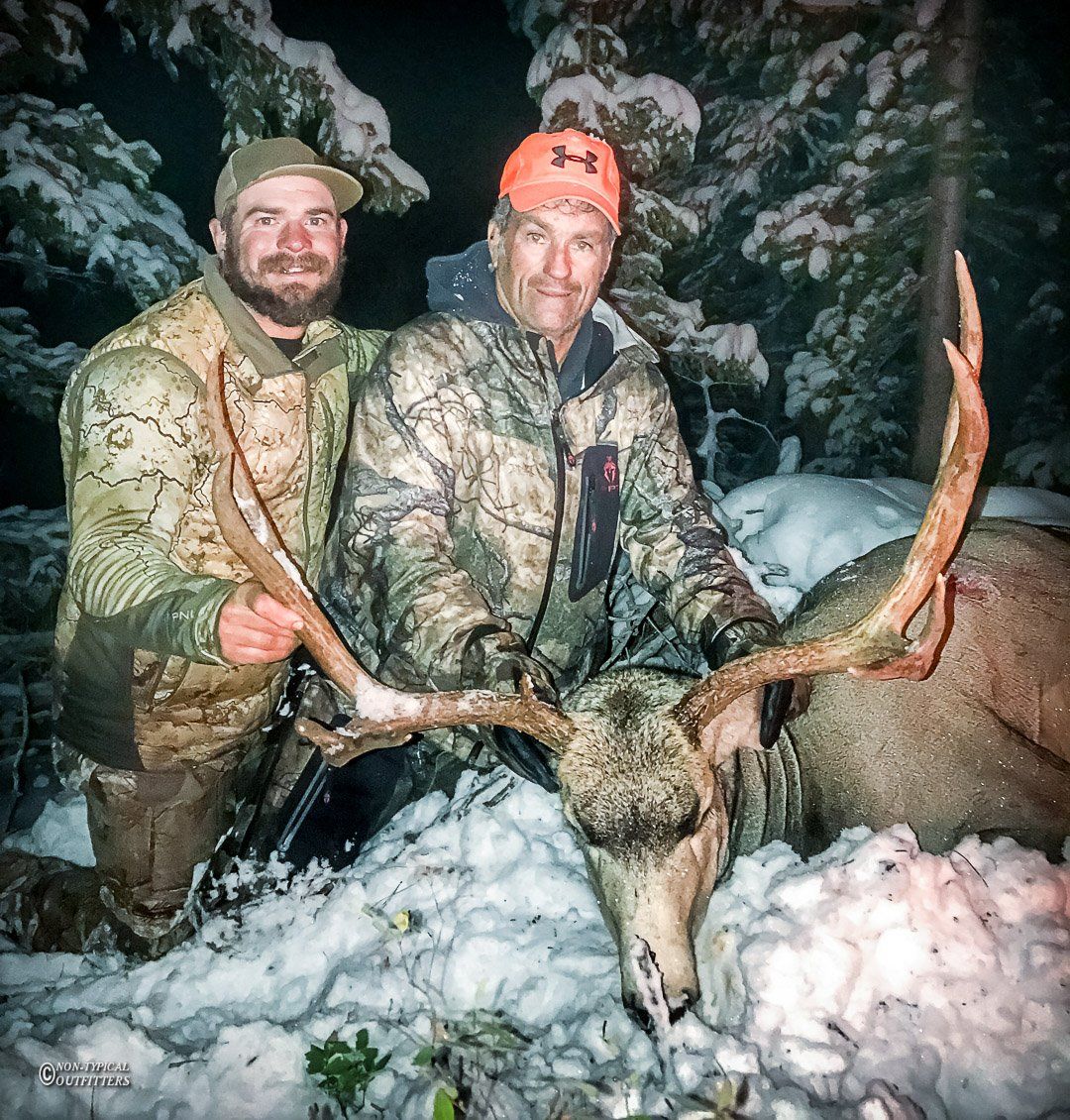 Two men pose with a large elk in the snow. They wear camo and the elk has large antlers.