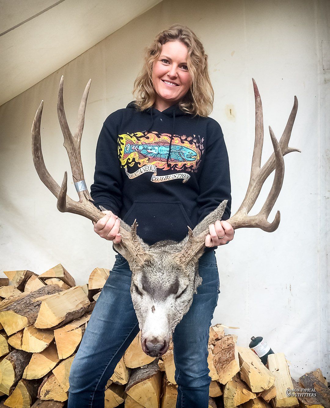 A woman in a black hoodie holds a deer head in front of a pile of logs