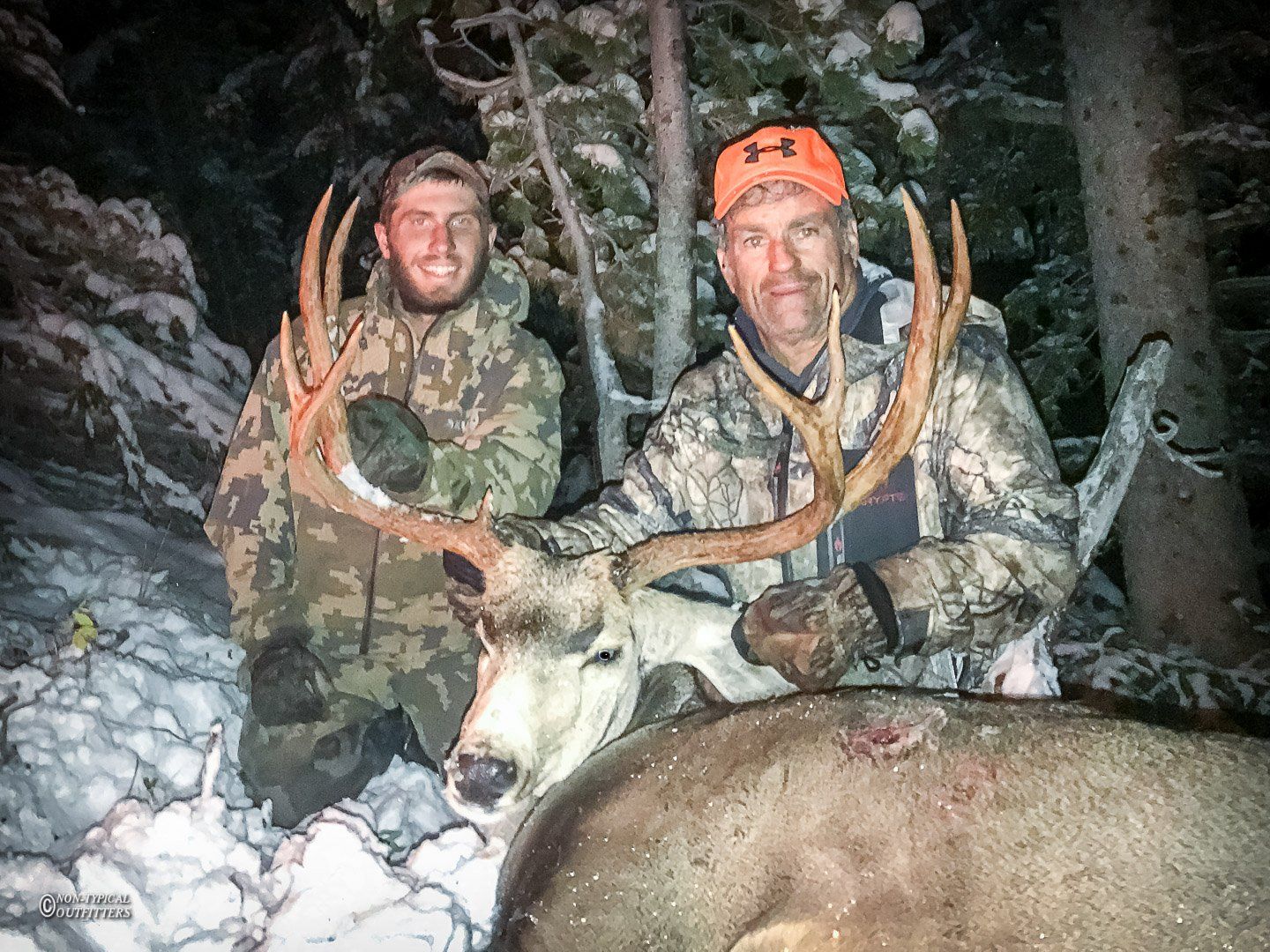 Two men pose with a large buck deer in snowy woods. One man wears an orange hat.