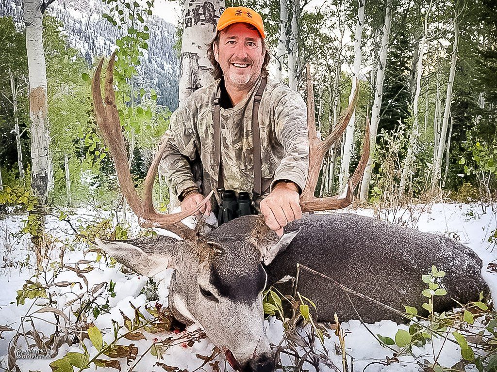 Man in camo and orange hat kneels beside a large buck on a snowy forest floor.