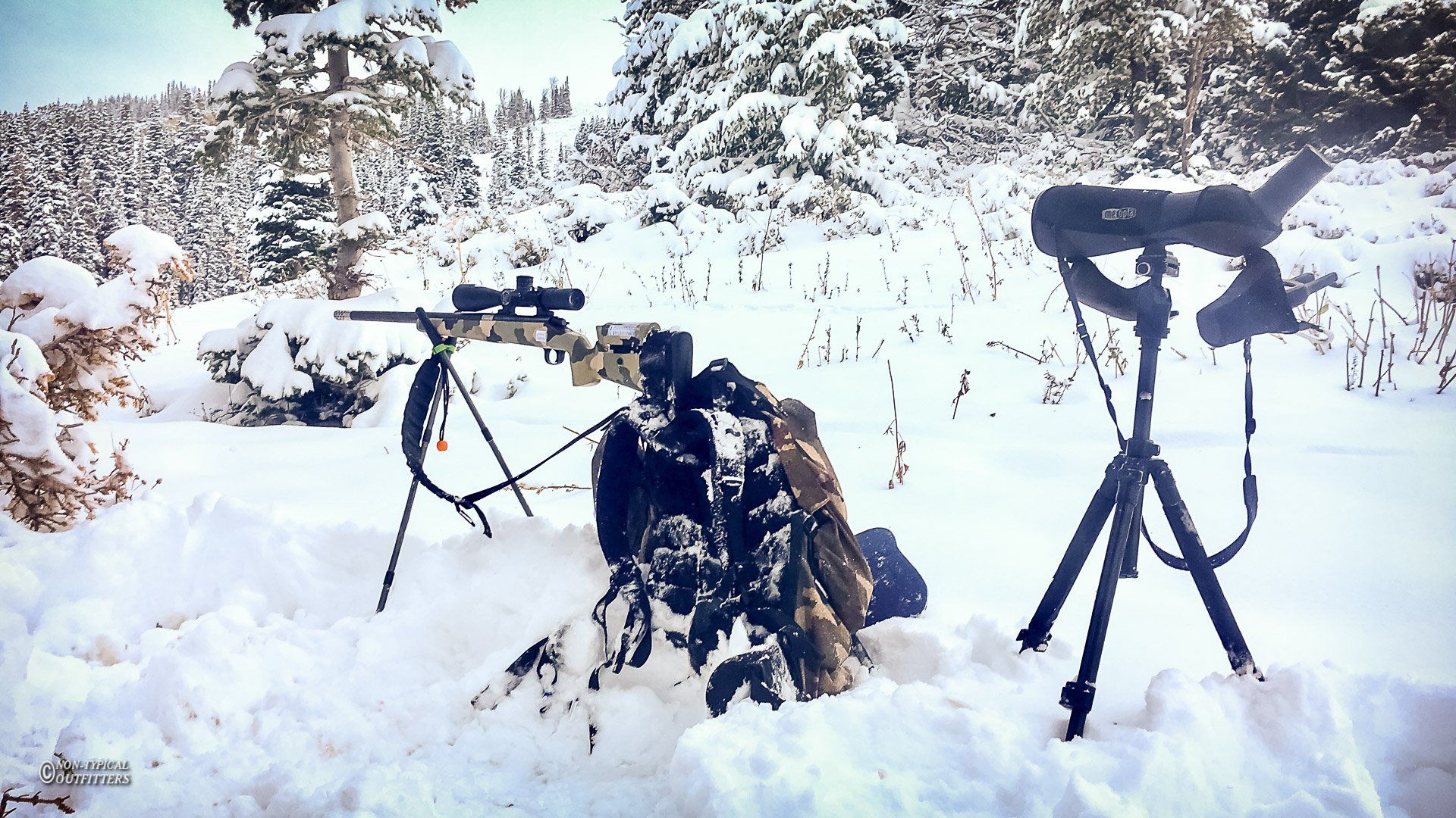 Snowy scene with a rifle on a tripod, spotting scope, and camouflage gear; winter landscape.