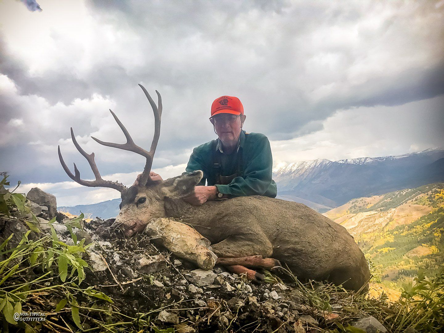 Hunter with a buck on a rocky mountainside. The deer has large antlers, and the man wears orange and green clothing.