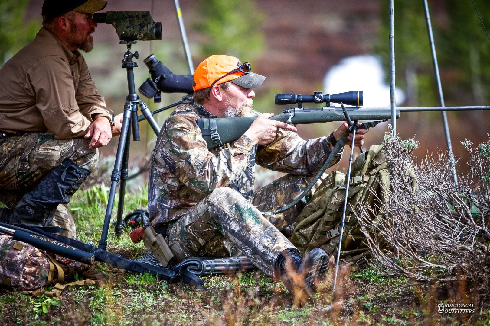 Two men in camouflage hunting gear aim rifles, one wearing an orange cap in a forest setting.