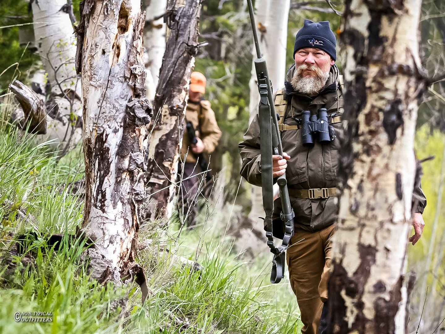 Man with rifle and binoculars in forest, another person in background.