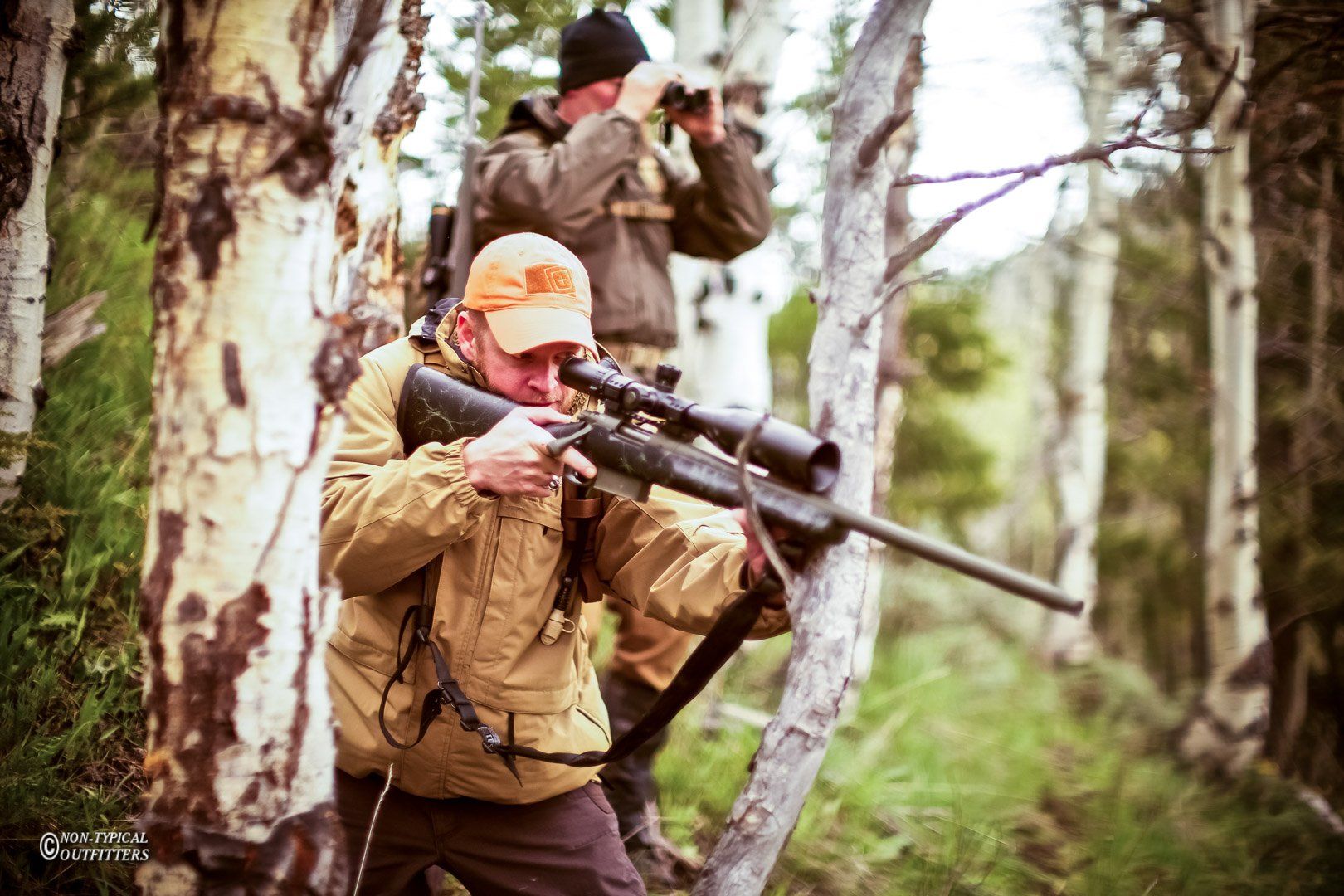 Two hunters in a forest: one aims a rifle, the other uses binoculars, surrounded by trees.