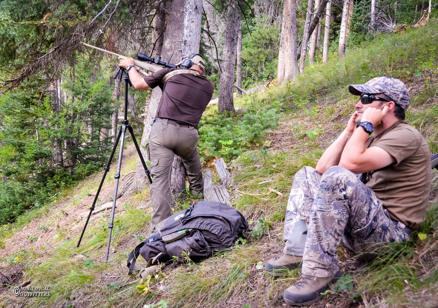 Two men hunting in a wooded area. One aims a rifle on a tripod, the other sits looking on.