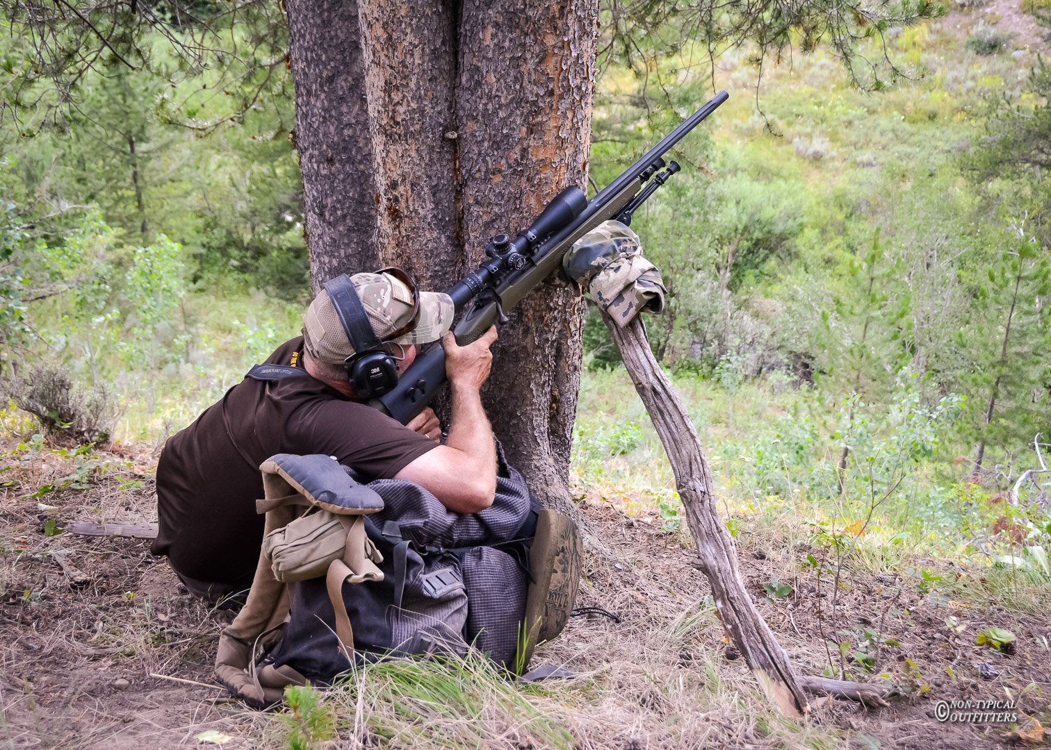 Man aims rifle, supported by a tree, in a forest setting; camouflage backpack nearby.