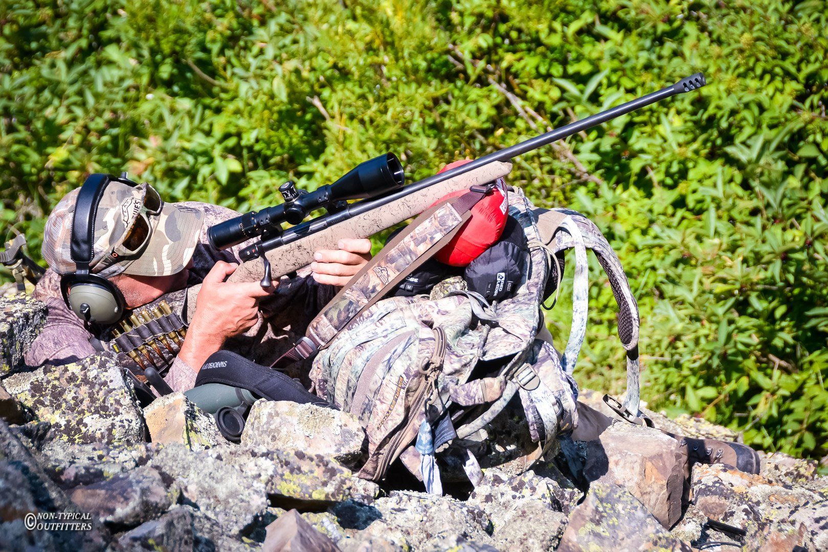 Hunter in camouflage aiming a rifle with a scope, nestled among rocks and foliage.