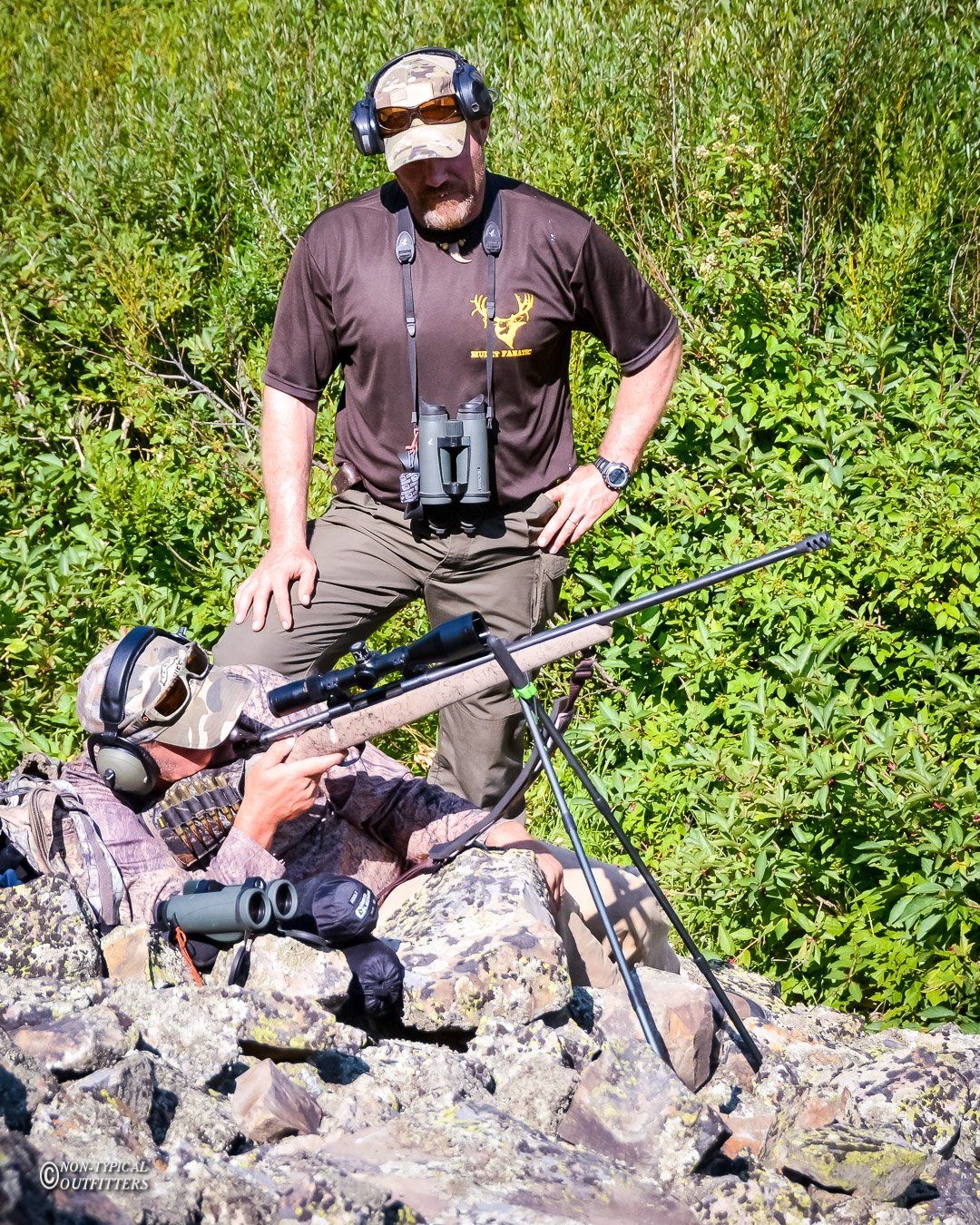 Two people, one prone with a rifle on a bipod, another standing, both in camouflage, in a wooded area.