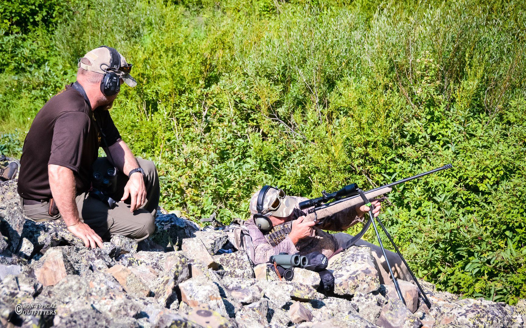 Man watches another man aiming a rifle in a grassy outdoor setting. Both wear ear protection.