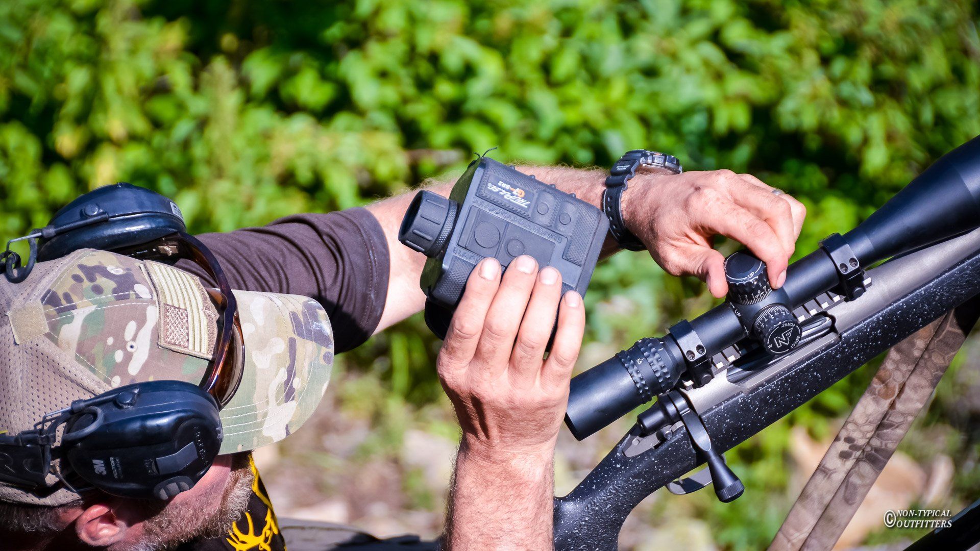 Person adjusting a black rangefinder on a rifle, outdoors with foliage in the background.