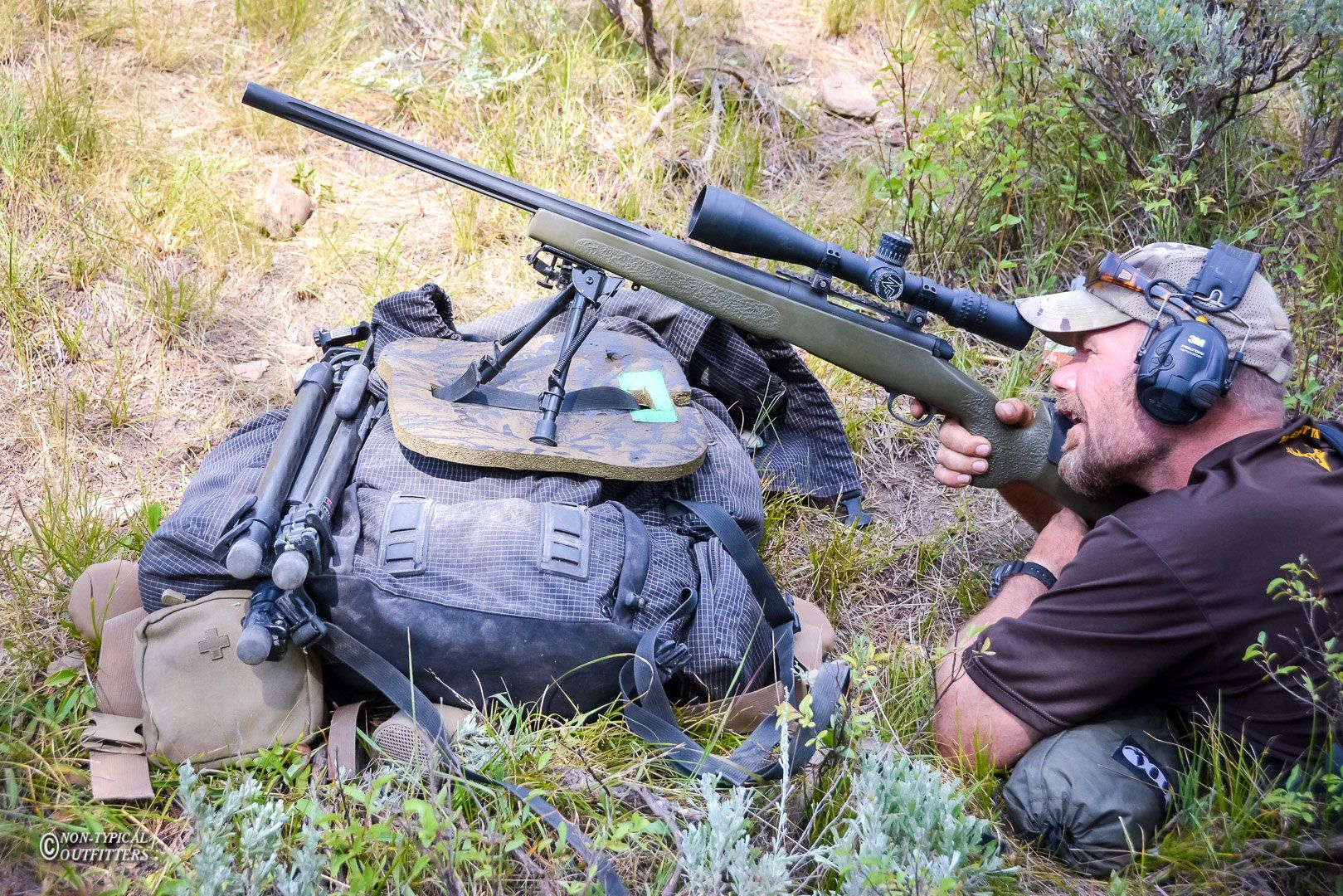 Man aiming a rifle over a camo backpack in a field. He's wearing ear protection and focused.