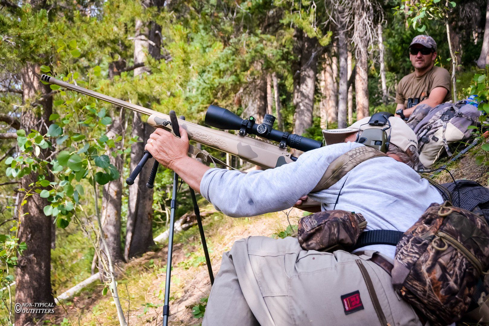 Hunter aiming rifle, supported by bipod, in forest. Another man watches.