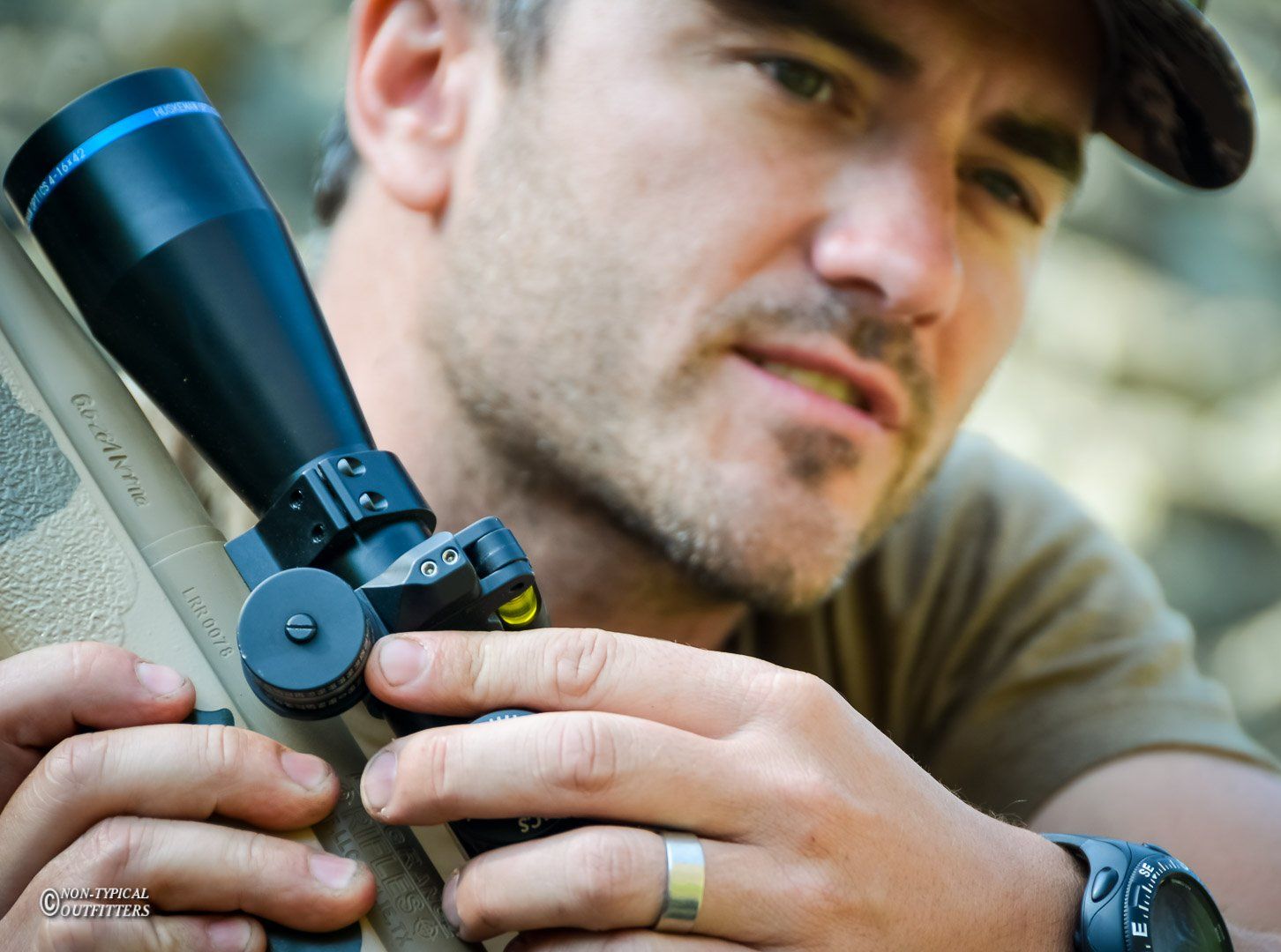 Man adjusting a rifle scope outdoors. He has a concerned expression and wears a watch.