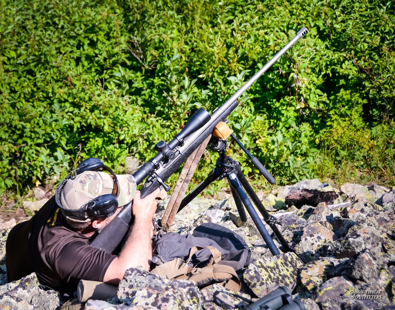 Person aiming rifle with scope on tripod, surrounded by rocks and foliage.