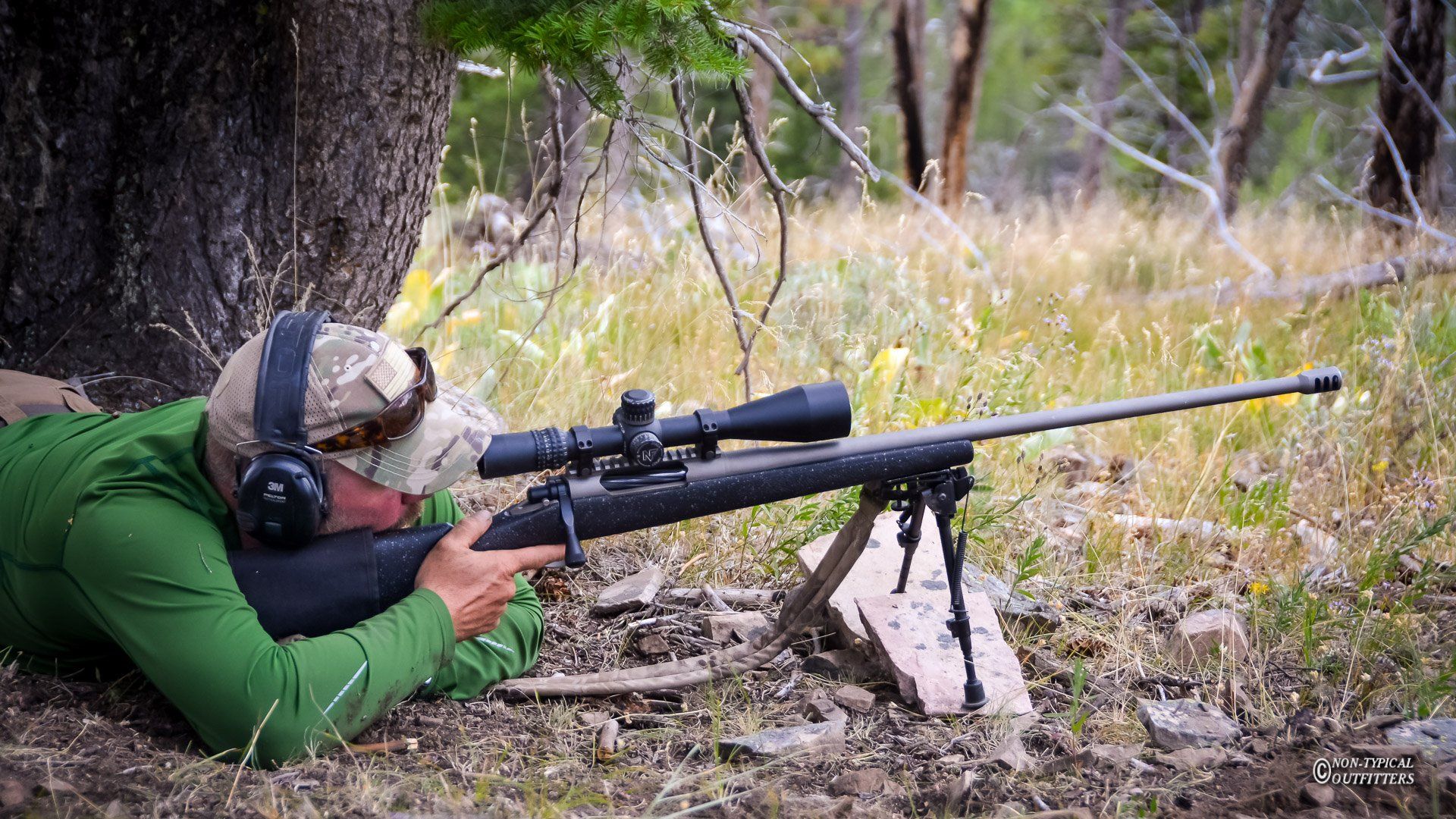 Person prone, aiming rifle with scope in a wooded area. Rifle is on a bipod. Wearing camo hat and ear protection.