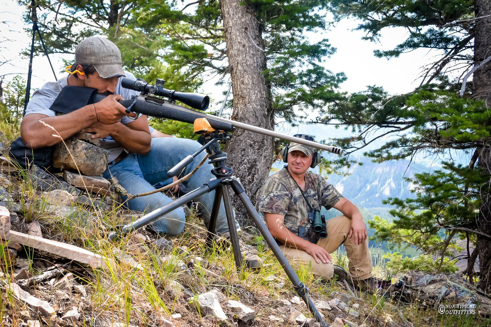 Two hunters with rifles on a hillside, one aiming, the other observing. Forest and mountains in the background.