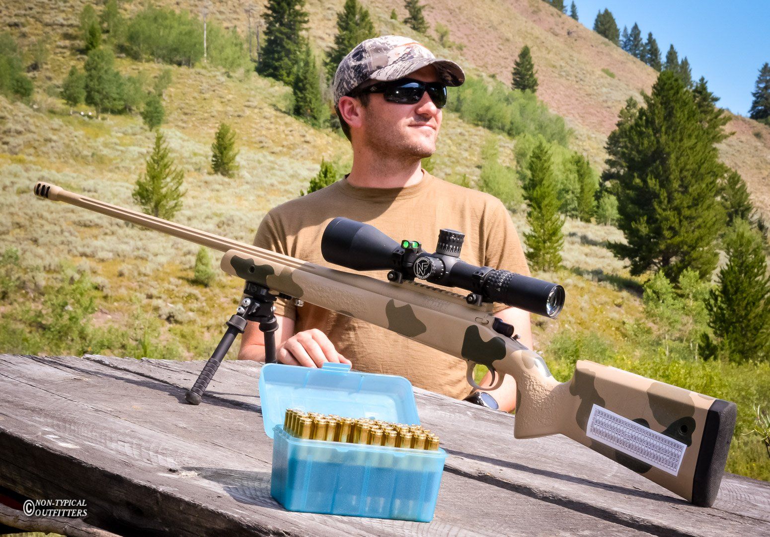 Man in sunglasses and camo hat with a rifle, next to ammo box, outdoors on a sunny day.
