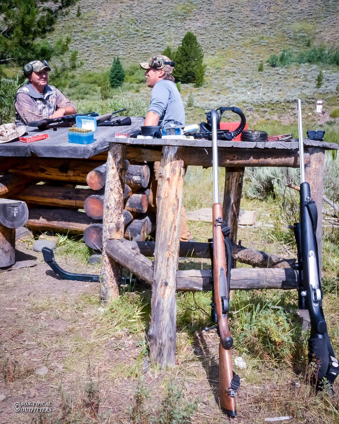 Two people at a wooden table with rifles. Ammunition and gear are visible. Outdoors, sunny day.