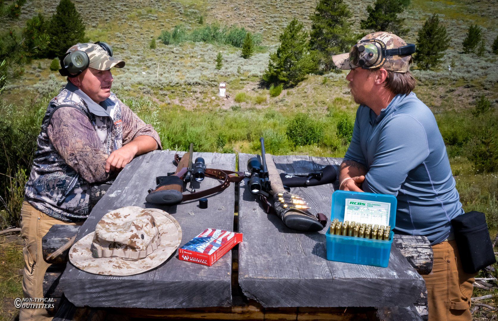 Two people at a shooting range, talking over rifles on a table with ammunition and targets in the background.
