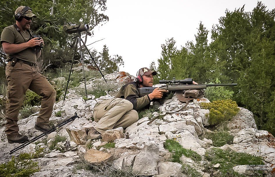 Two men on a rocky hillside; one aims a rifle, the other uses binoculars. Overcast sky.