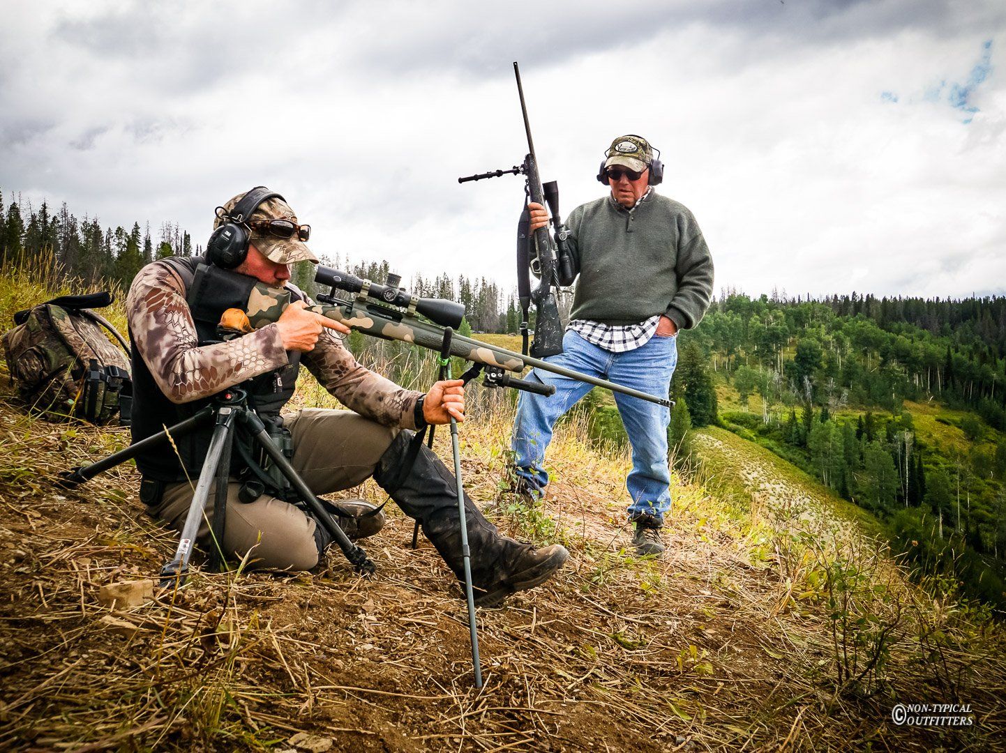 Man aiming rifle on a tripod, another man watches on a hillside, overcast sky, green trees.