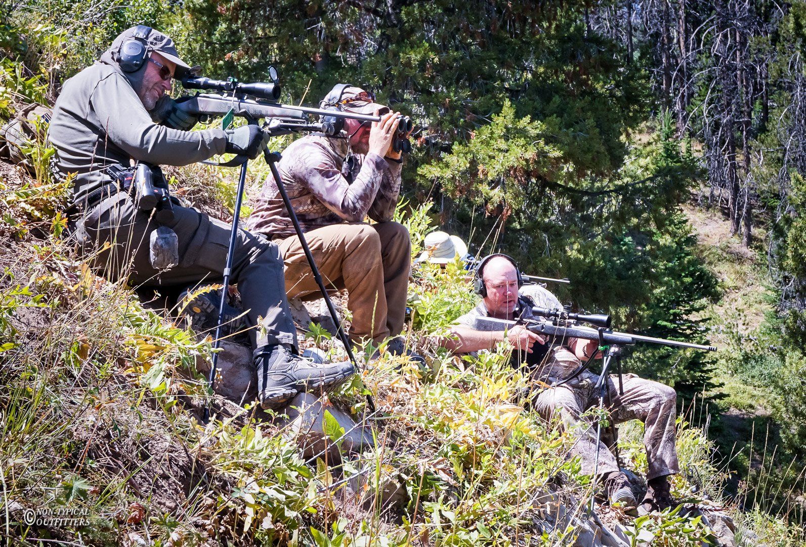Three people in camouflage gear hunting in a wooded area, aiming rifles and using binoculars.