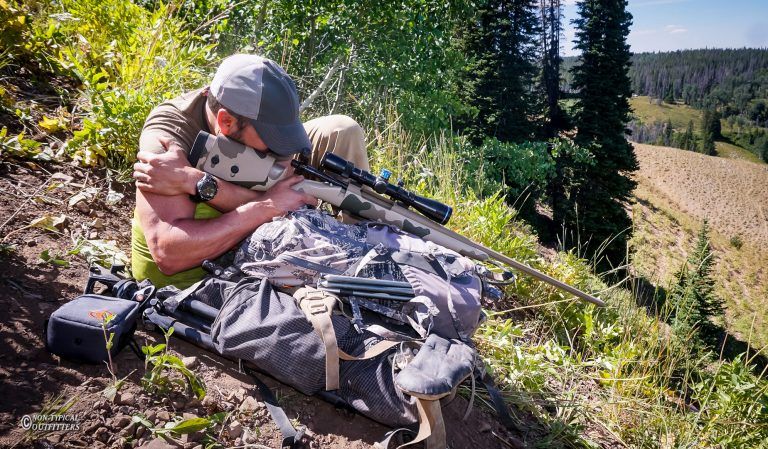 Man aiming a rifle in a mountainous area. He is leaning against a backpack on a grassy slope with trees in the background.
