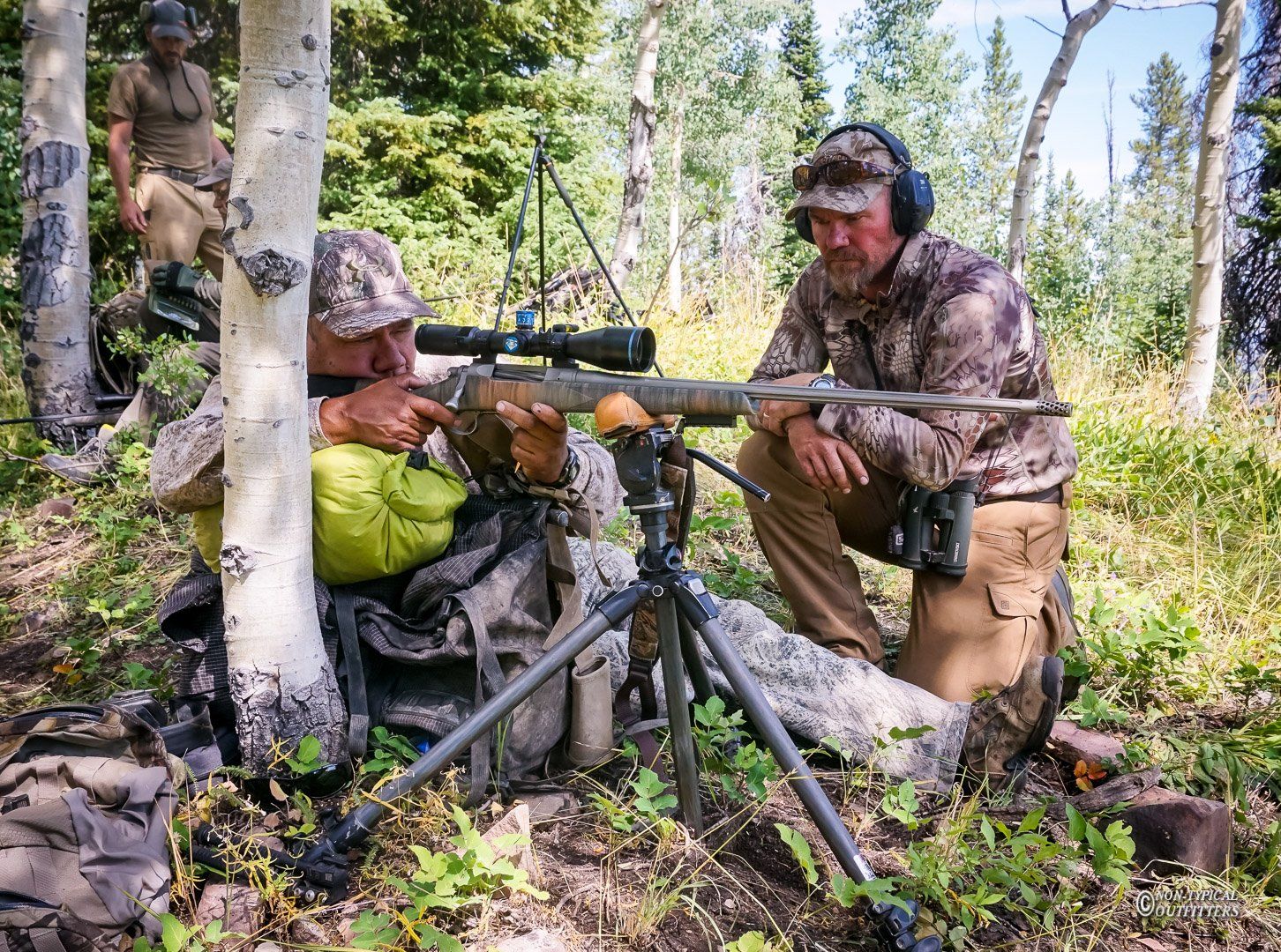 Two men in camouflage gear hunting in a forest. One aims a rifle supported by a tripod; the other kneels, observing.
