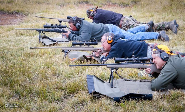 Four people prone, aiming rifles at a target in a grassy field. All wear ear protection.